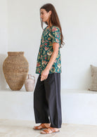 Woman stands sideways wearing a relaxed fit bottle green based floral print top against a white wall.