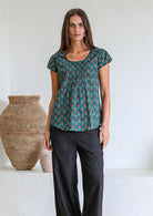 Woman wears bottle green based floral 100t% cotton cap sleeve top with pin tuck detail across the bust and a scoop neckline