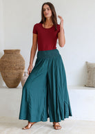 Woman wearing a maroon top and teal wide leg pants standing in a light filled room