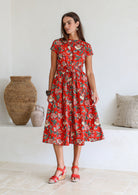 Woman wears red based floral printed cotton button through cotton dress with hidden side pockets, short sleeves and a detachable waist tie.
