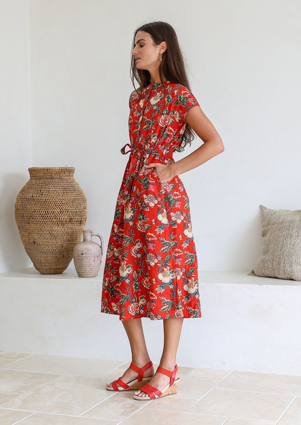 Woman wearing a red floral 100% cotton dress with one hand in pocket standing in a room with a white wall and decorative items.