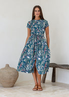 Model stands wearing a retro style midi length cotton dress with button through front, fabric waist tie and short sleeves.