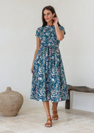 Model standing wearing a 100% cotton button through midi length dress with teal based floral print.
