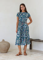 Woman wears 100% cotton button through midi length dress with short sleeves and pockets in floral print on a teal base.
