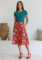 Woman wearing a basic green top and red floral a-line cotton skirt with side zipper and one hand in pocket.