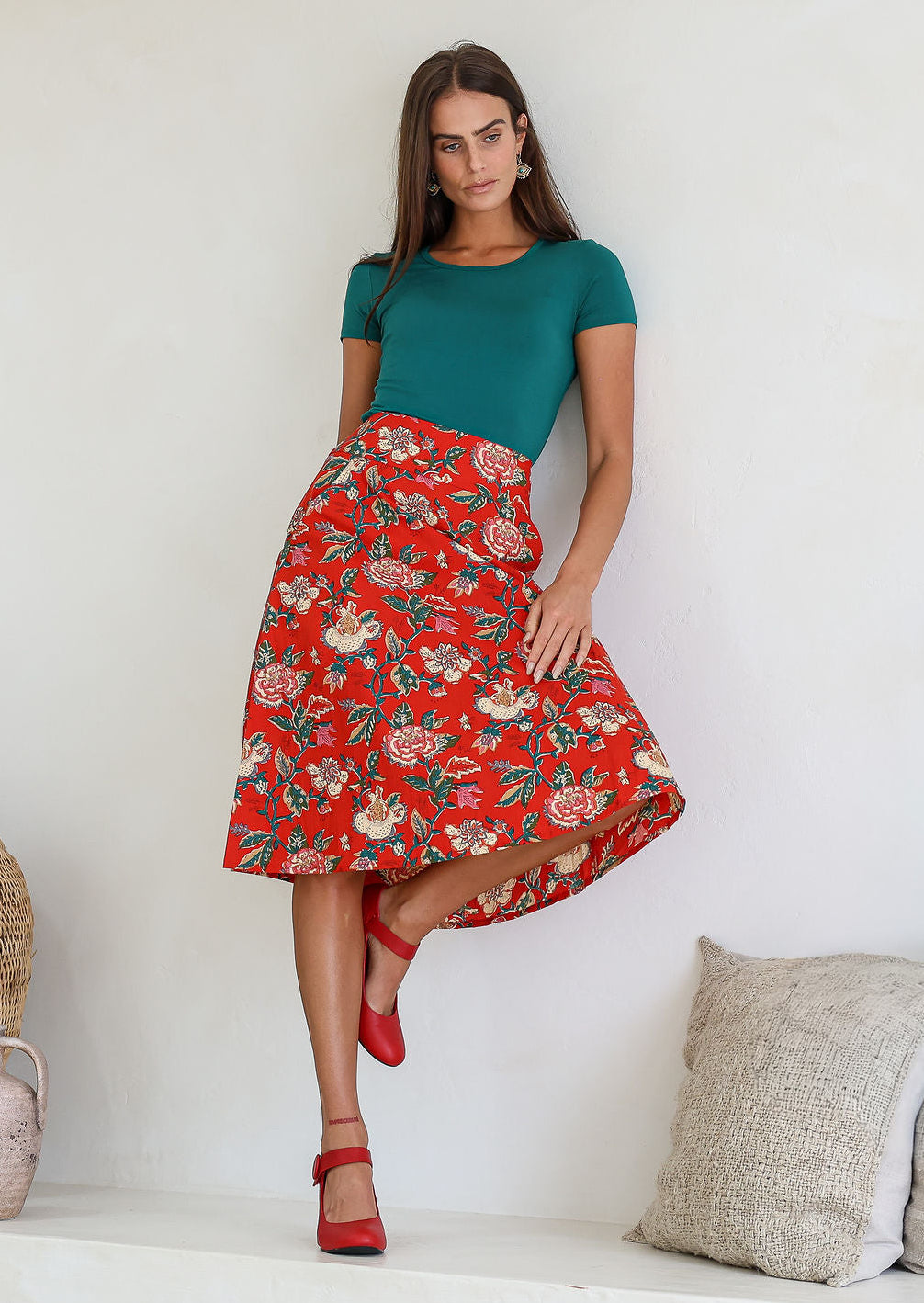 woman leans on wall wearing a knee length floral cotton skirt with a-line shape, pockets and side zipper.