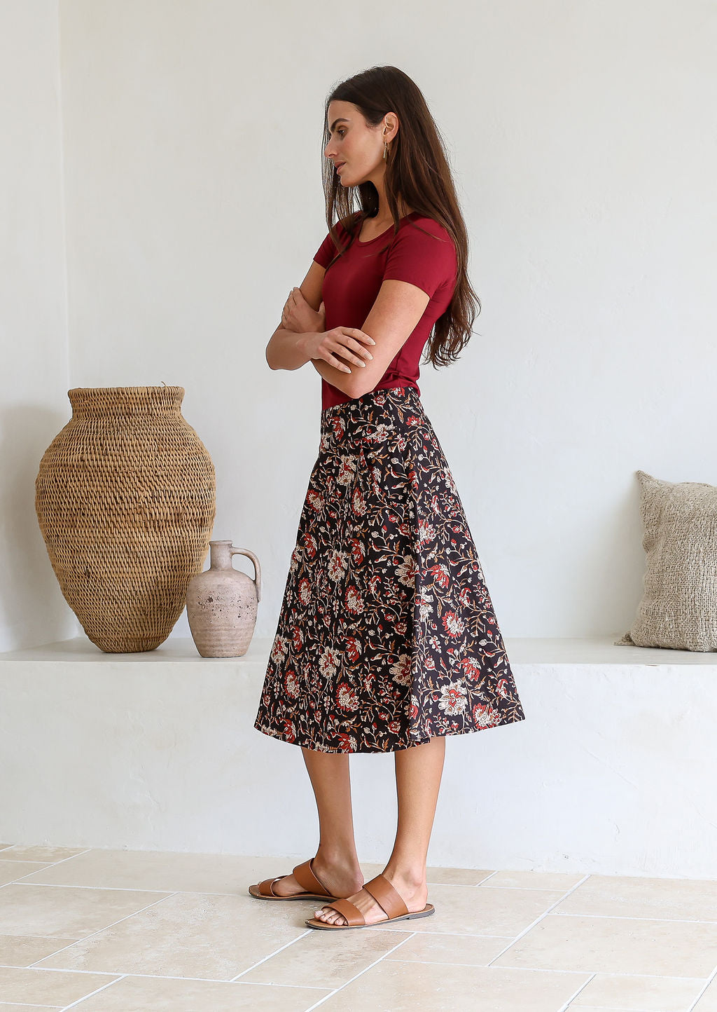 Woman stands sideways in a knee length floral cotton skirt with a-line shape, pockets and side zipper.