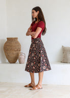 Woman stands sideways in a knee length floral cotton skirt with a-line shape, pockets and side zipper.