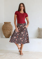 Woman wearing a maroon top and floral  cotton skirt standing in a room with a woven vase and pillows.