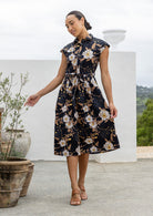 Model stands with wearing a midi length floral printed shirt style cotton dress with cap sleeves, buttoned bodice and a removable waist tie
