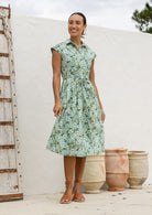 Woman wearing a midi length sage green  based floral cotton dress with buttoned bodice, standing in front of a white wall with rustic elements.