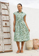 Model stands with wearing a midi length floral printed shirt style cotton dress with cap sleeves, buttoned bodice and a removable waist tie
