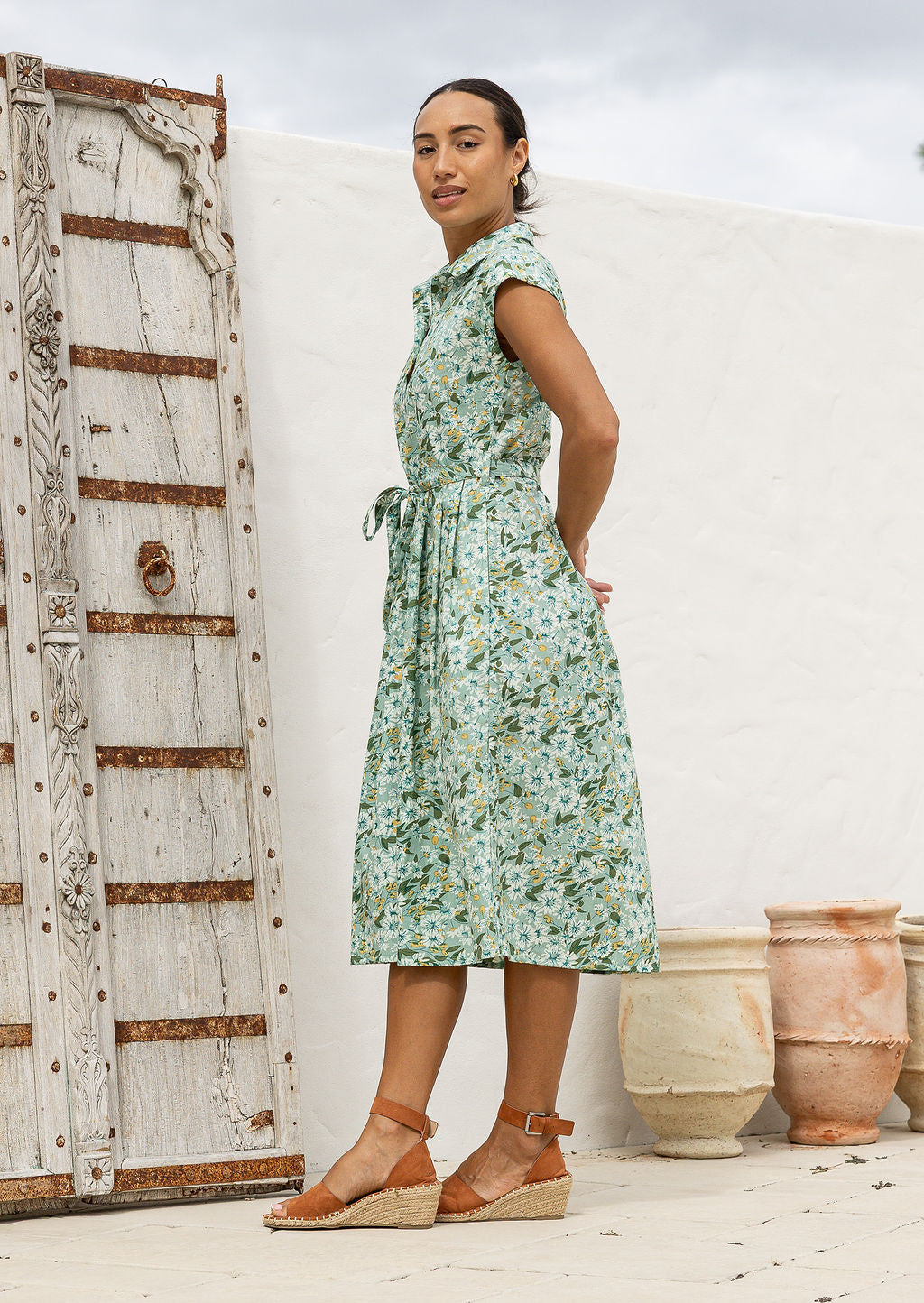 Model stands sideways near wearing a mid length, shirt style floral print cotton dress with sage green based floral print.