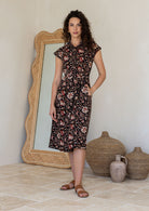 Woman wearing a midi length black based floral cotton dress with buttoned bodice, standing against a white wall and decorative vases
