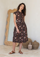 Model stands with wearing a midi length floral printed shirt style cotton dress with cap sleeves, buttoned bodice and a removable waist tie