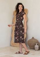 Model stands sideways near ceramic pots, wearing a midlength, shirt style Indian traditional  floral print cotton dress with with rust, tan and sand tones on a black base.