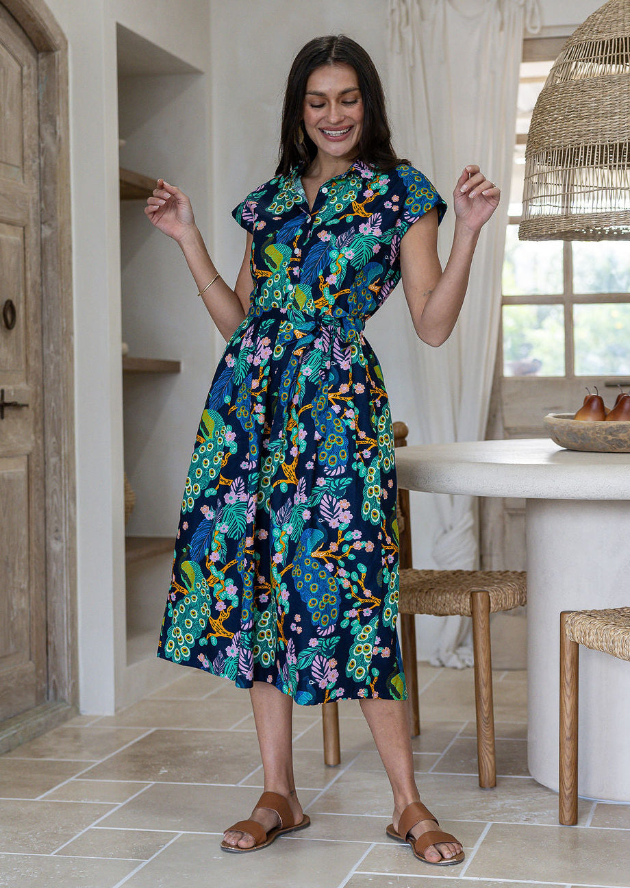 A woman modelling a cap-sleeve cotton midi dress with a vibrant floral and peacock print in teal, green, blue, yellow, and pink on a dark navy background, set in a bright, modern rustic kitchen.