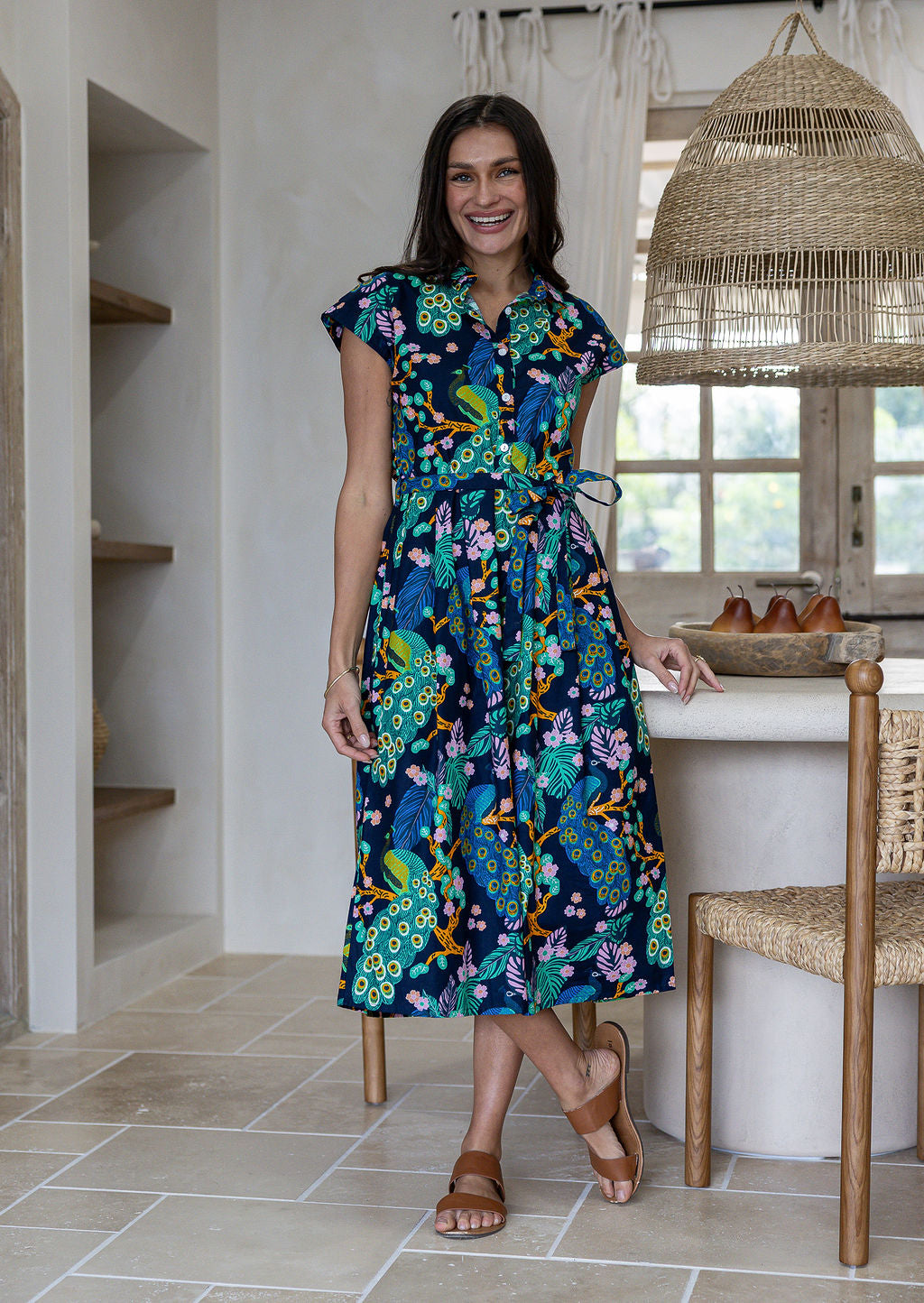 A woman modelling a cap-sleeve cotton midi dress with a collar, buttoned bodice and waist tie, in a vibrant multicolour floral and peacock print