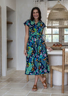 A woman modelling a cap-sleeve cotton midi dress with a collar, buttoned bodice and waist tie, in a vibrant multicolour floral and peacock print