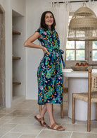 A woman modelling a cap-sleeve cotton midi dress with a colourful floral and peacock print on a dark navy base, with hand on hip