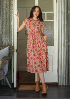 Woman wearing a midi length cream  based floral cotton dress with buttoned bodice, standing in front of a white door.