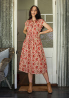 Model stands with wearing a midi length floral printed shirt style cotton dress with cap sleeves, buttoned bodice and a removable waist tie