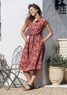 Woman leans on a wall wearing a shirt style cotton midi length dress with buttoned bodice, pockets, collar and cap sleeves