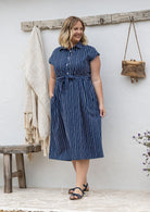A woman wearing a navy white vertically striped midi 100% cotton dress with one hand in pocket