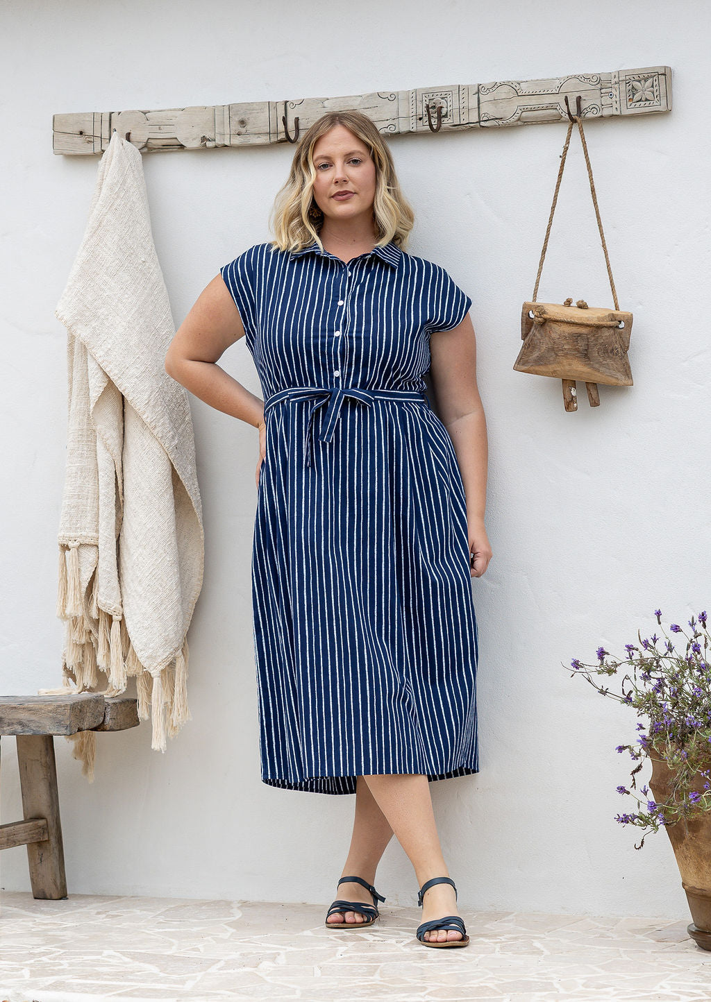 Model stands with wearing a midi length floral printed shirt style cotton dress with cap sleeves, buttoned bodice and a removable waist tie