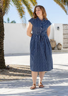 Model stands with wearing a midi length floral printed shirt style cotton dress with cap sleeves, buttoned bodice and a removable waist tie