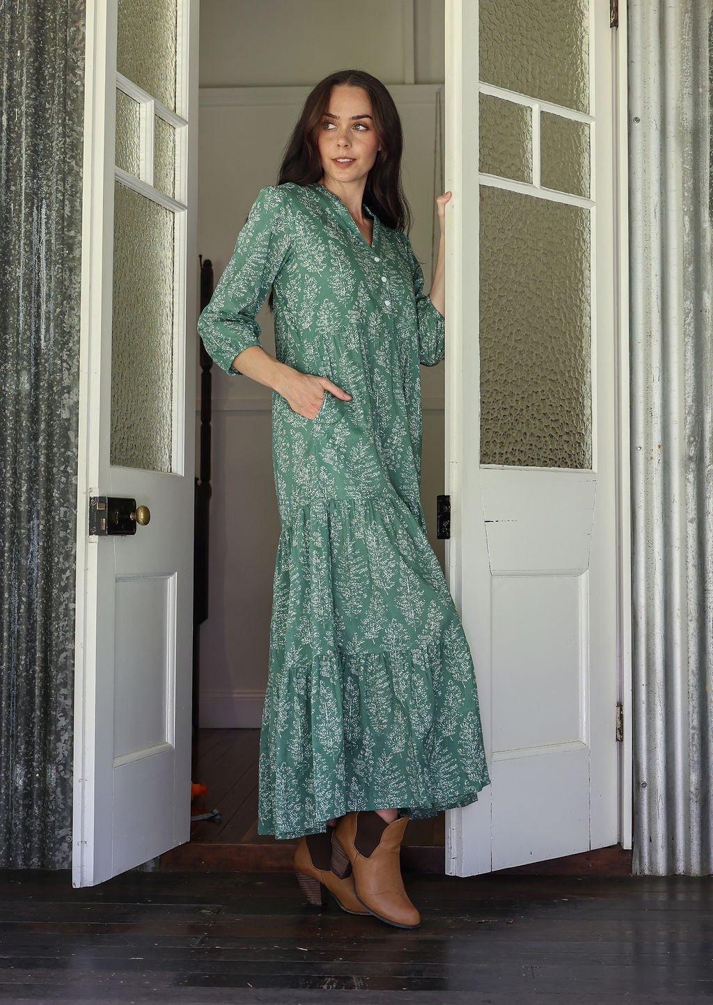 Woman stands side ways wearing a 100% cotton tiered maxi dress in sage green based floral print with one hand in pocket