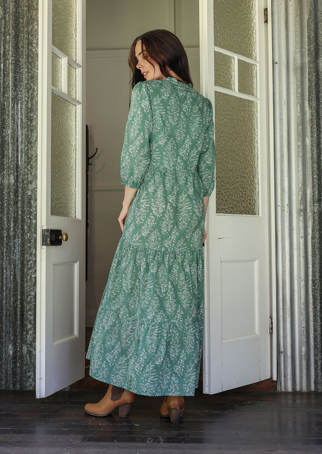 Woman stands backwards wearing a 100% cotton tiered maxi dress in cream based floral print with 3/4 sleeves, pockets and mandarin collar