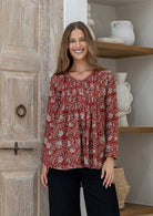Woman wearing a red based floral cotton long sleeve shirt with pin tucks across the back of the bodice, the back of shirt is longer than front