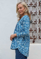 Woman wearing a blue floral button through cotton top with long sleeves, pleats at the front and back, and a longer hem at the back.