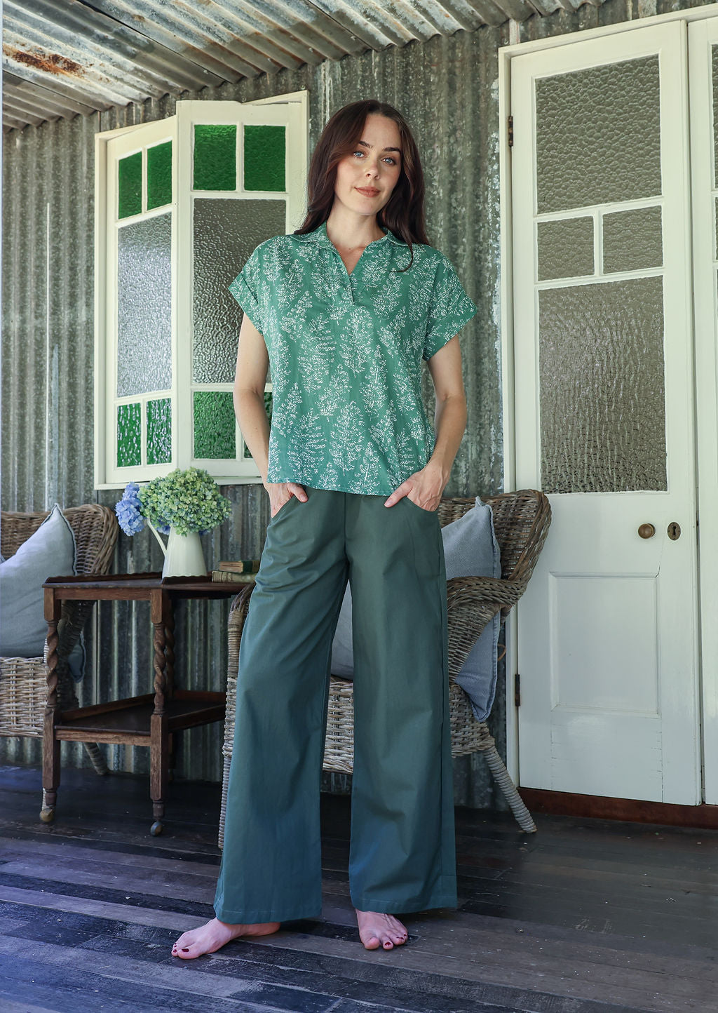 Woman wearing a sage green based floral 100% cotton top and plain pants standing in a rustic indoor setting.