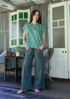 Woman wearing a sage green based floral 100% cotton top and plain pants standing in a rustic indoor setting.