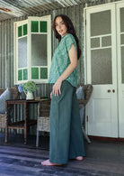 Woman stands sideways wearing a sage green based floral cotton top with short folded cuff sleeves, collar and V neckline