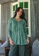 Woman wearing a sage green based cotton floral long sleeve relaxed fit blouse with a V-neckline, standing in a room