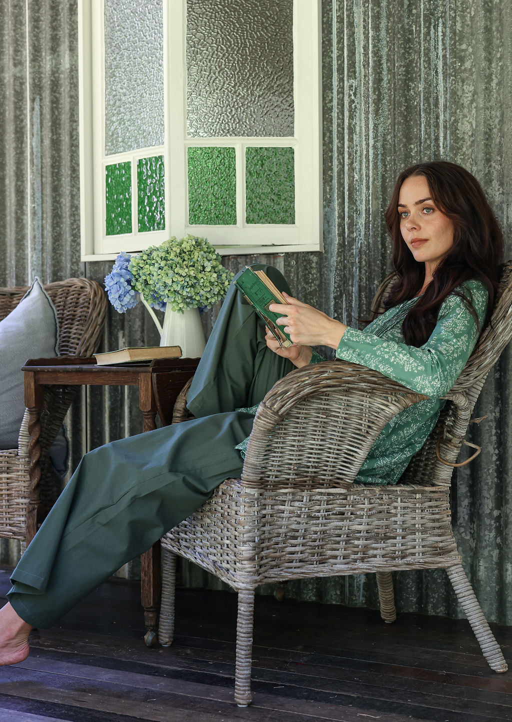 Woman sitting in a wicker chair reading a book in a room with textured walls and a window.
