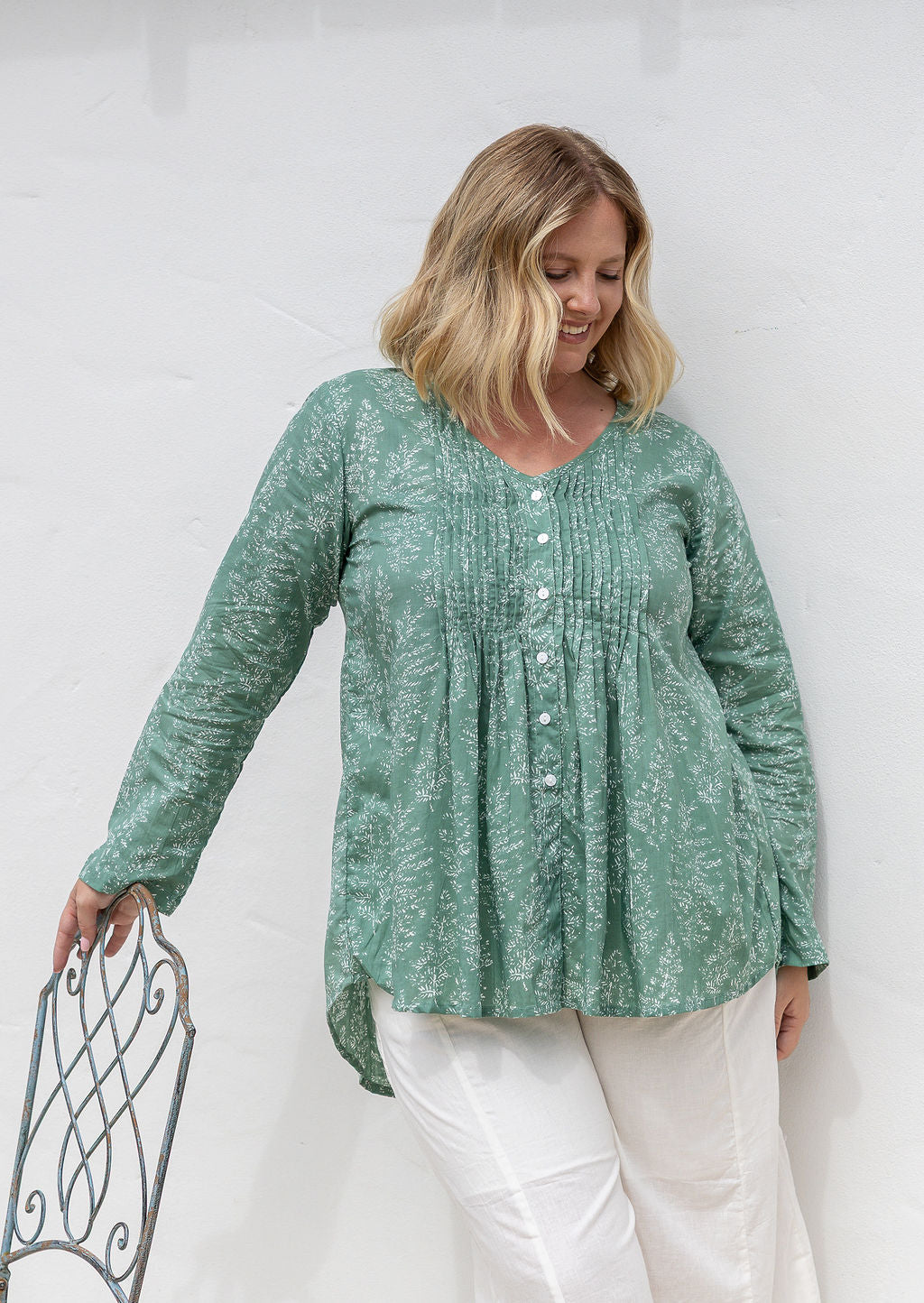Woman wearing a sage green based floral cotton long sleeve shirt with pin tucks across the back of the bodice, the back of shirt is longer than front