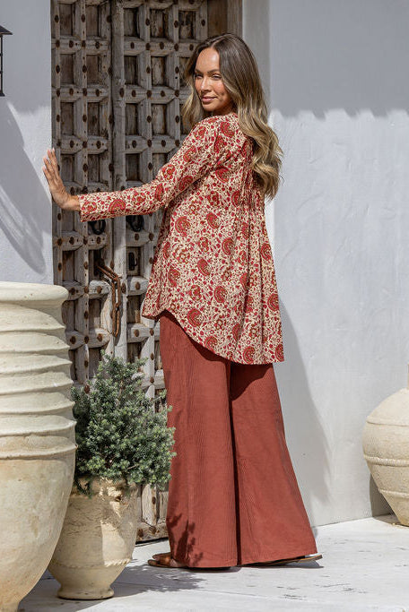 Woman in a red floral blouse and rust-colored pants standing against a textured wall.