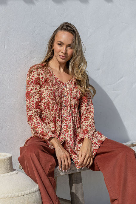 Woman sitting on a stone block wearing a patterned blouse and red pants against a light gray wall.