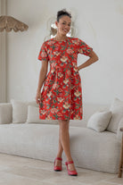 Woman wearing a relaxed fit, above knee length retro floral cotton dress with vibrant red backdrop accented by elegant multicoloured floral vines.