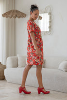 Woman stands sideways wearing a relaxed fit, red based floral cotton print dress with pockets and empire waistline, standing in a living room.