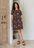 Model wears short sleeved, above knee 100 percent cotton dress with high round neckline and one hand in pocket.