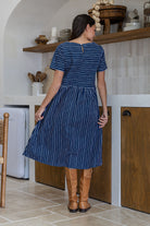 Woman stands backwards wearing a 100% cotton relaxed fit short sleeve dress in navy based white striped print. 