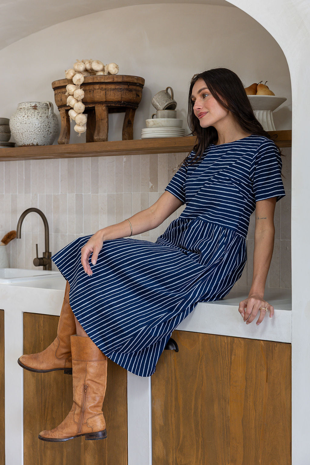 A woman wearing a navy blue white striped short-sleeve midi dress sits on a white counter in a rustic kitchen setting. 
