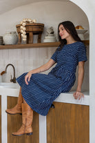 A woman wearing a navy blue white striped short-sleeve midi dress sits on a white counter in a rustic kitchen setting. 