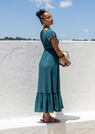 Side view of Woman in a teal maxi, perfect Christmas dress holding a ceramic pot against a white wall with blue sky.
