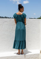 Back view of Woman in a teal maxi, perfect Christmas dress holding a ceramic pot against a white wall with blue sky.
